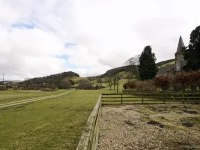 a field with a fence and a church and a road at The Coach House - Hw7782 in Llanarmon Dyffryn-Ceiriog