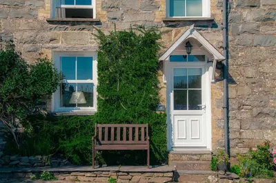 a wooden bench in front of a stone house with a white door at Little Cott - Cosy Welsh Cottage, a hiking retreat in Maentwrog