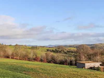 a wooden building in a field with trees and hills at Meadow Lodge in Corwen